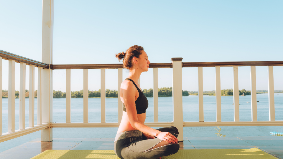 A woman practicing mindfulness and meditation on a balcony overlooking a serene lake, focusing on managing ADHD symptoms (tCaPC).